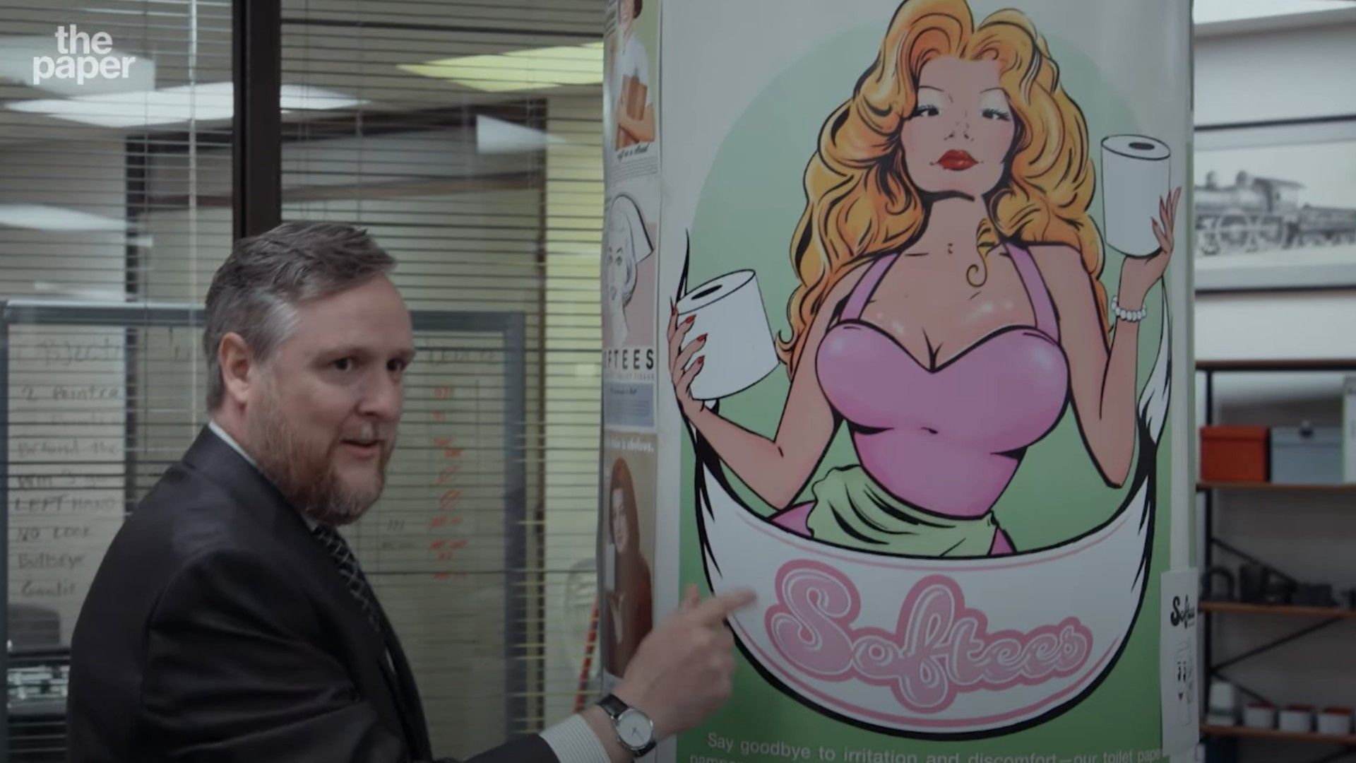 Un bărbat în costum lângă un poster cu o femeie blondă desenată care ține suluri de hârtie igienică