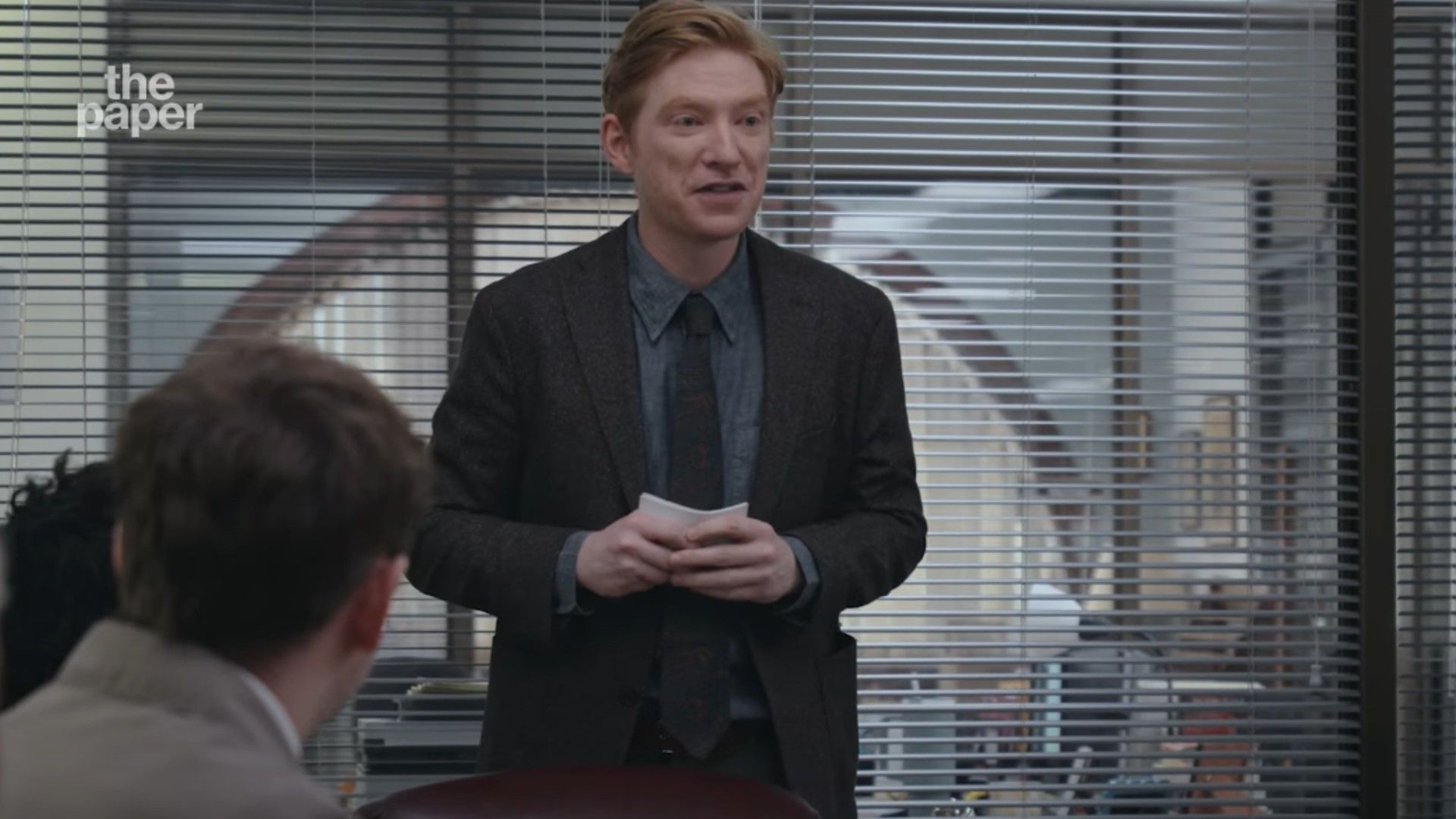 Domhnall Gleeson în costum stând într-o sală de conferințe