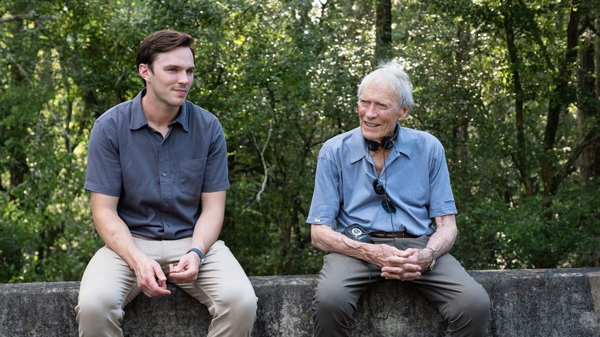 Nicholas Hoult și Clint Eastwood în haine casual stând pe o barieră de beton și vorbind cu o pădure în fundal