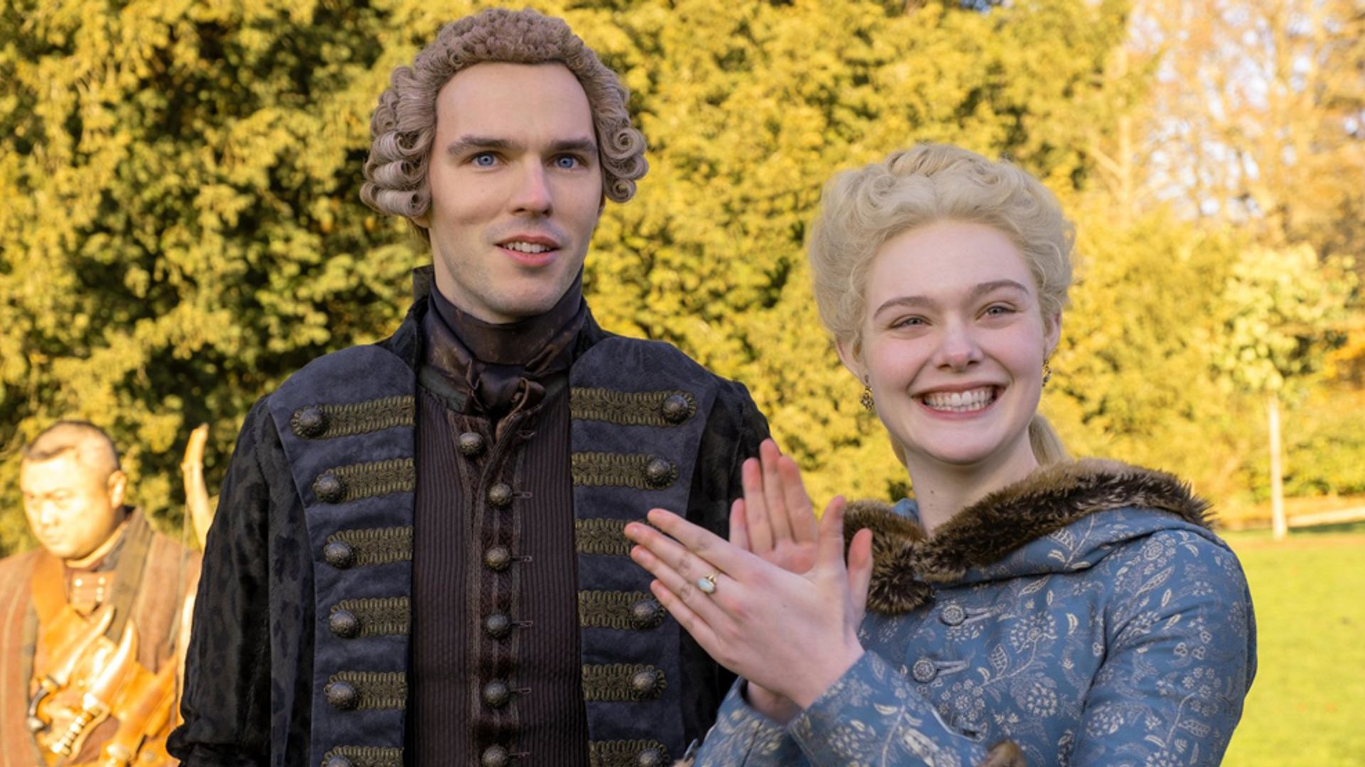 Nicholas Hoult și Elle Fanning în costume de epocă din secolul al XVIII-lea stând pe stradă, actrița zâmbește și bate din palme