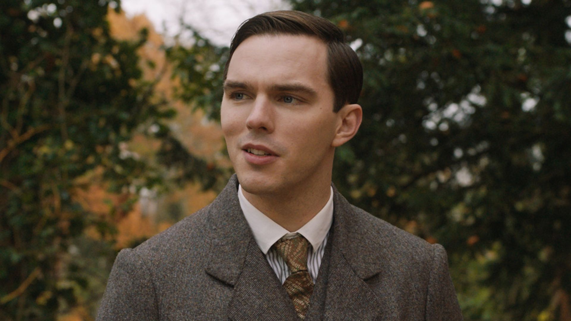 Nicholas Hoult în costum maro și cravată stând pe stradă în rolul tânărului J.R.R. Tolkien