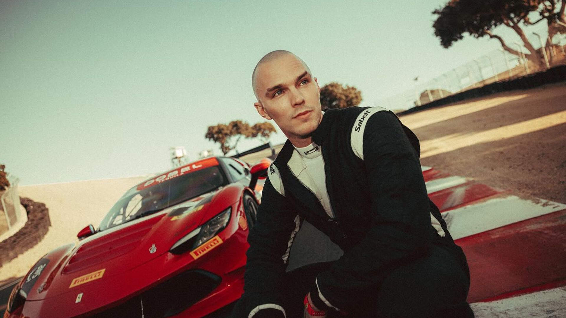 Nicholas Hoult în costum de curse stă pe pistă cu o mașină sport Ferrari roșie în spatele lui