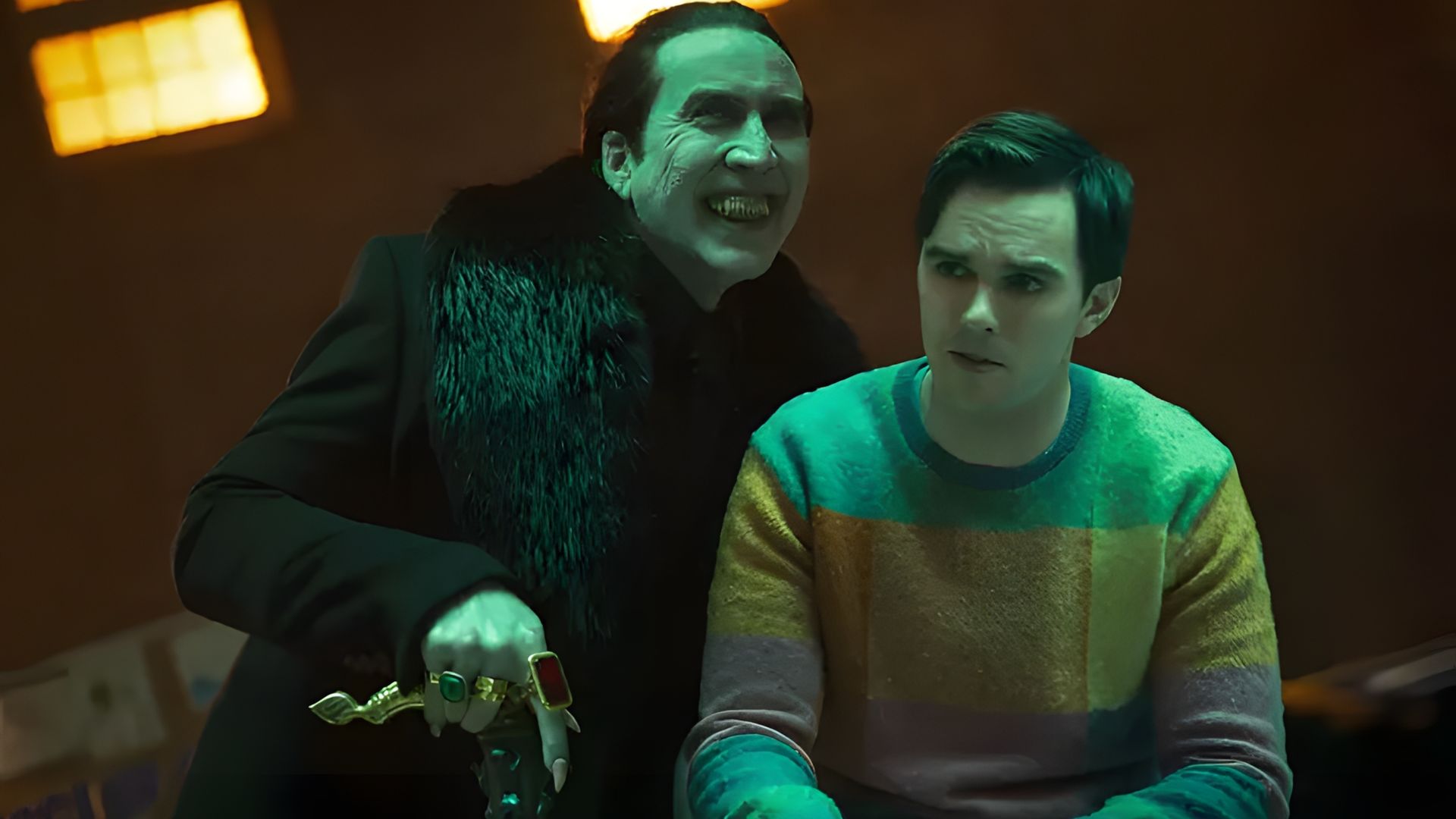 Nicolas Cage în haine negre cu inele planează amenințător peste Nicholas Hoult într-un pulover colorat