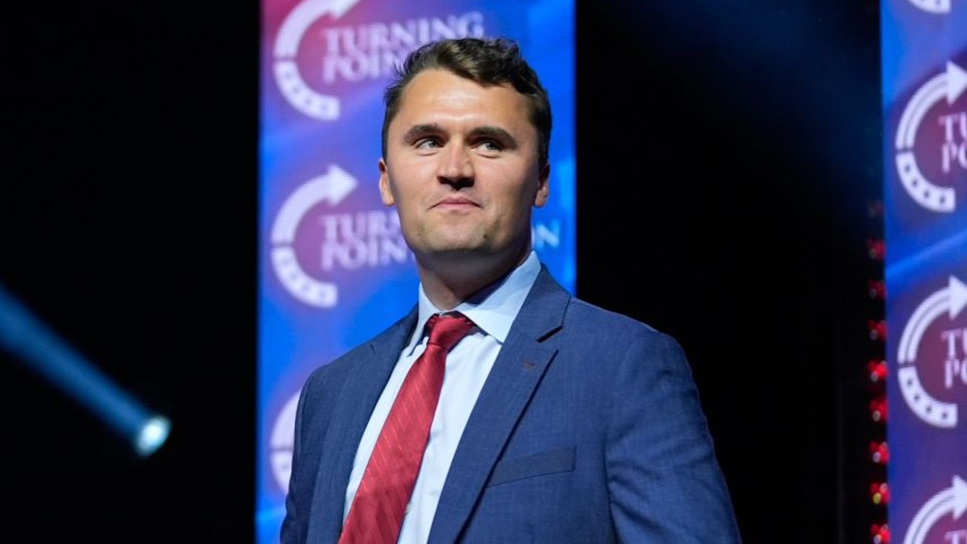 Charlie Kirk stă mândru pe un fundal întunecat, purtând un costum albastru