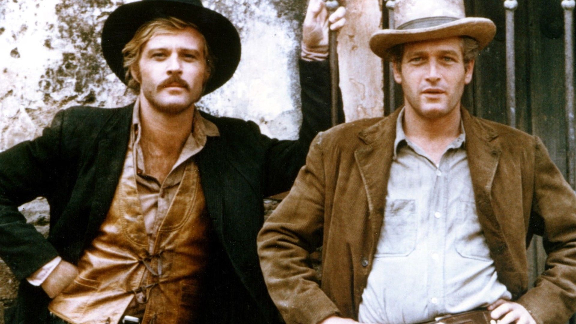 Robert Redford și Paul Newman cu pălării de cowboy și jachete, stând unul lângă altul lângă un perete, pozând pentru o fotografie