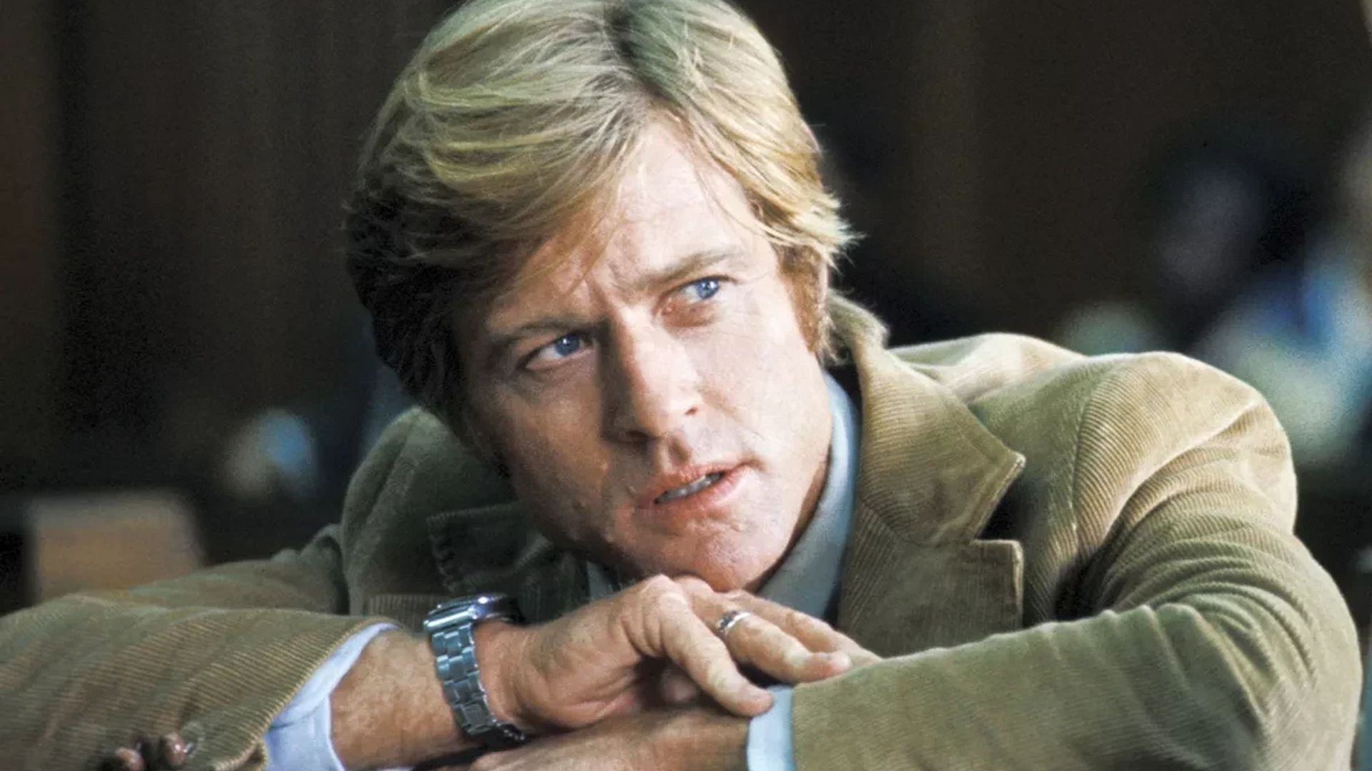 Robert Redford într-o jachetă maro cu un ceas pe încheietură se reazămă pe mâini și privește într-o parte