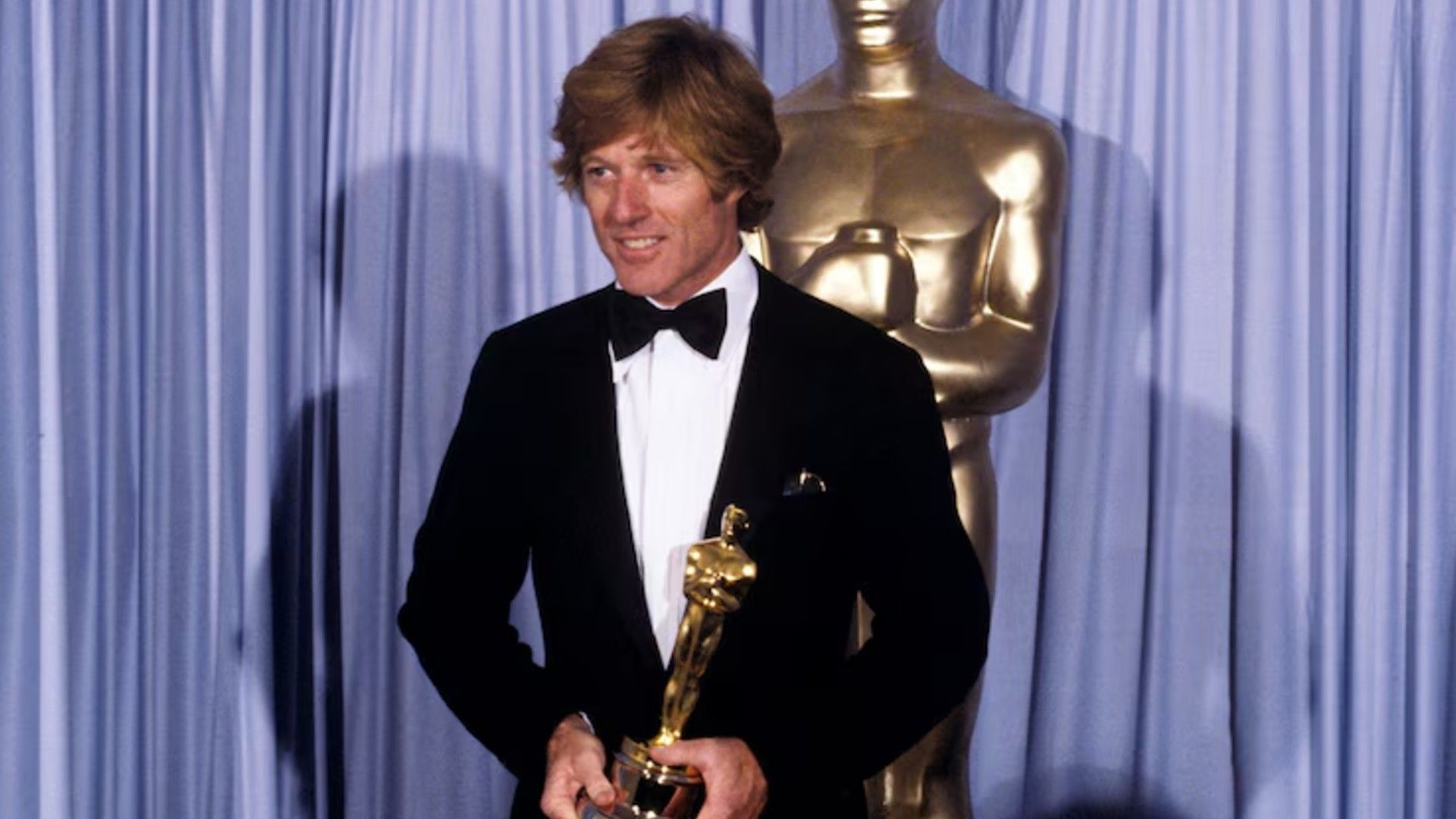 Robert Redford în costum stă pe scenă ținând un Oscar