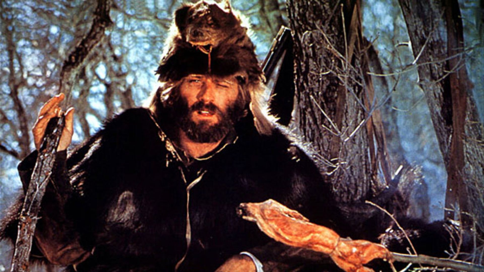 Robert Redford în îmbrăcăminte de blană și o căciulă din cap de animal preparând carne la foc de tabără în pădure, scenă din filmul "Jeremiah Johnson"