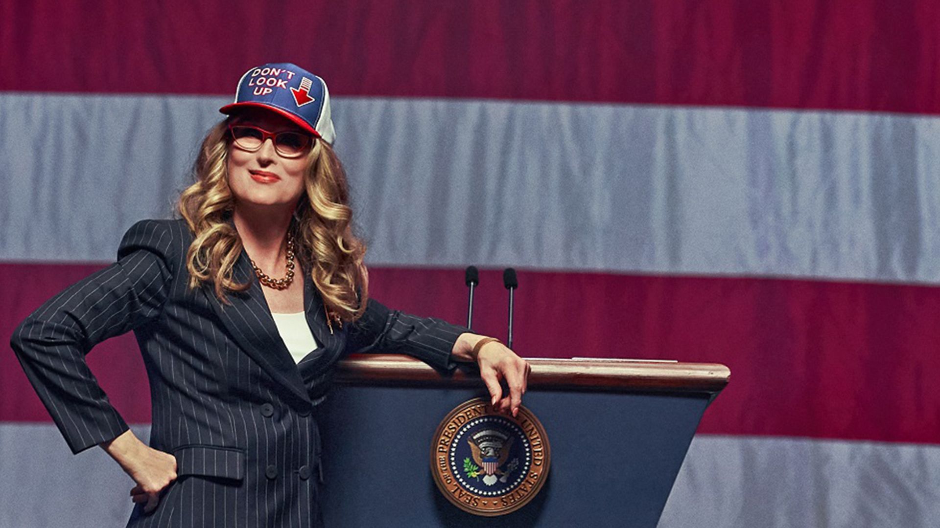 Meryl Streep în costum și șapcă cu steagul american stă pe fundalul steagului SUA, rezemată de o tribună