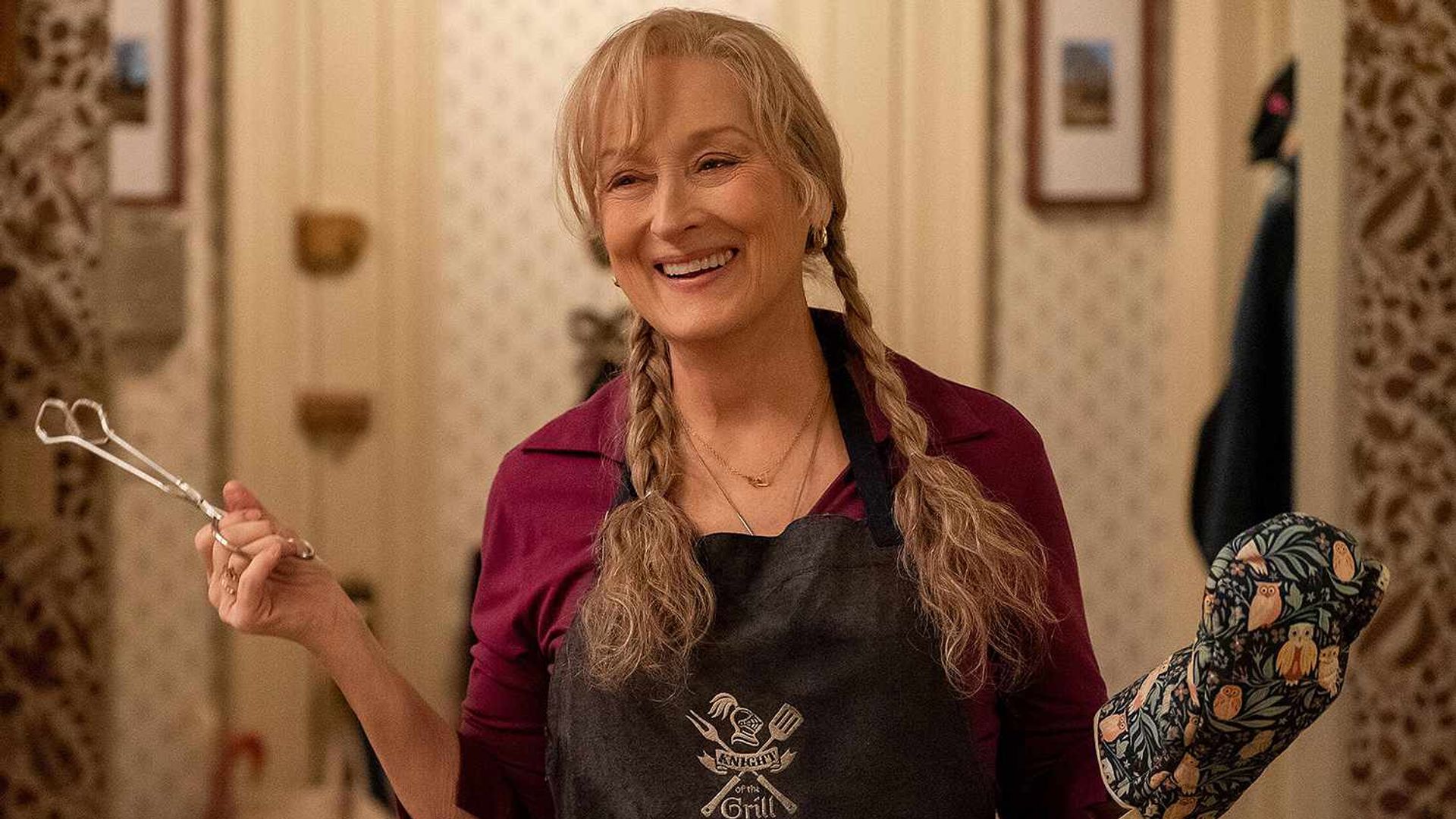 Meryl Streep cu împletituri ține o cleștă într-o mână, purtând o mănușă de bucătărie pe cealaltă