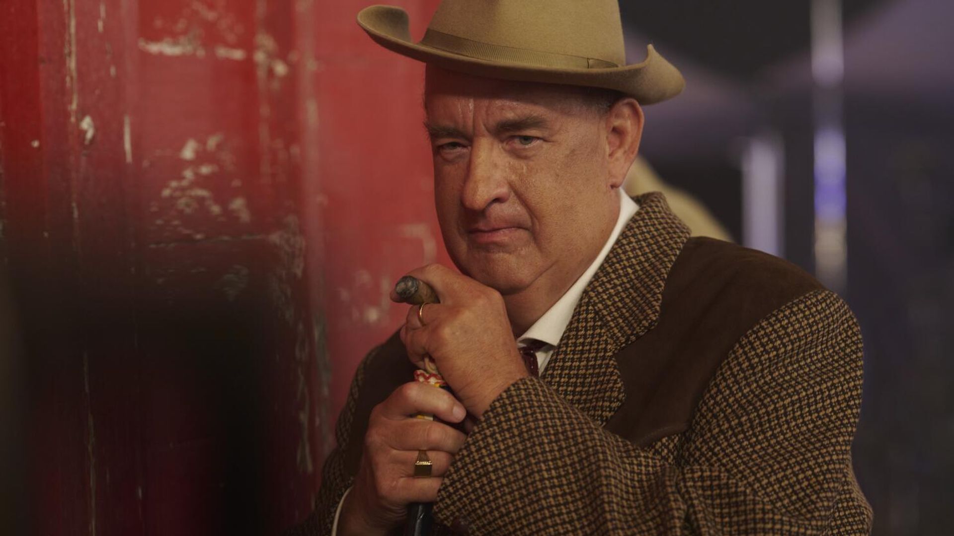 Tom Hanks în costum maro și pălărie se sprijină pe o bastonă, privind serios către cameră