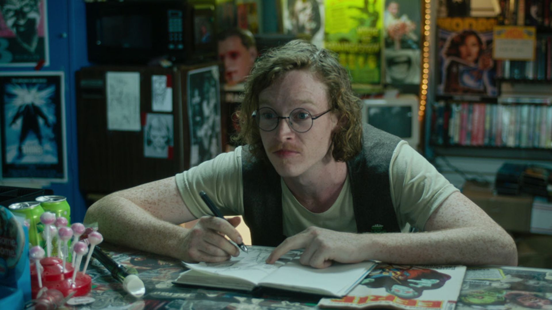 Caleb Landry Jones purtând ochelari și o vestă stă la o masă cu un carnet, desenând și privind într-o parte