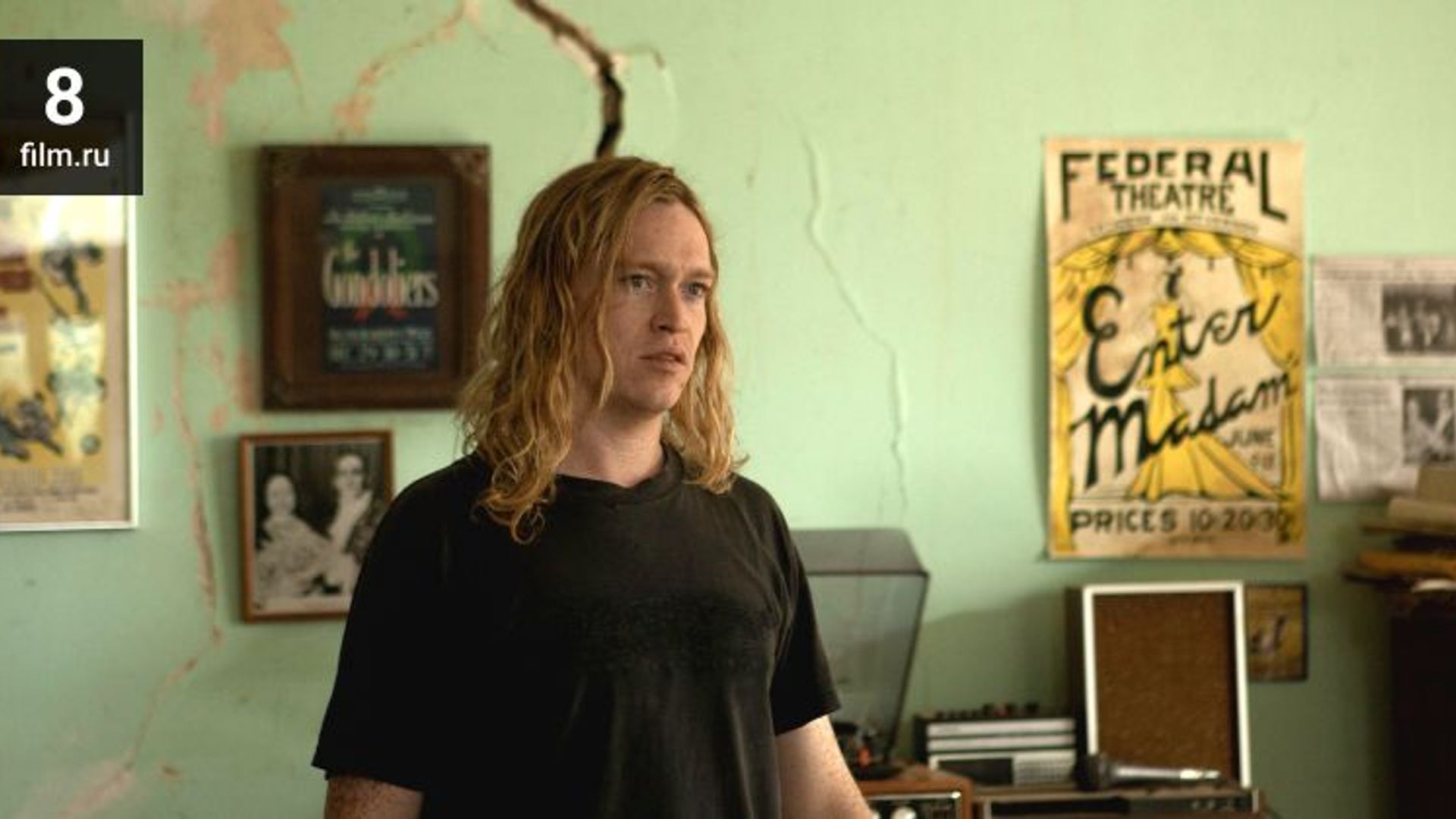 Caleb Landry Jones cu părul blond lung, îmbrăcat într-un tricou negru, stă într-o cameră cu postere pe perete