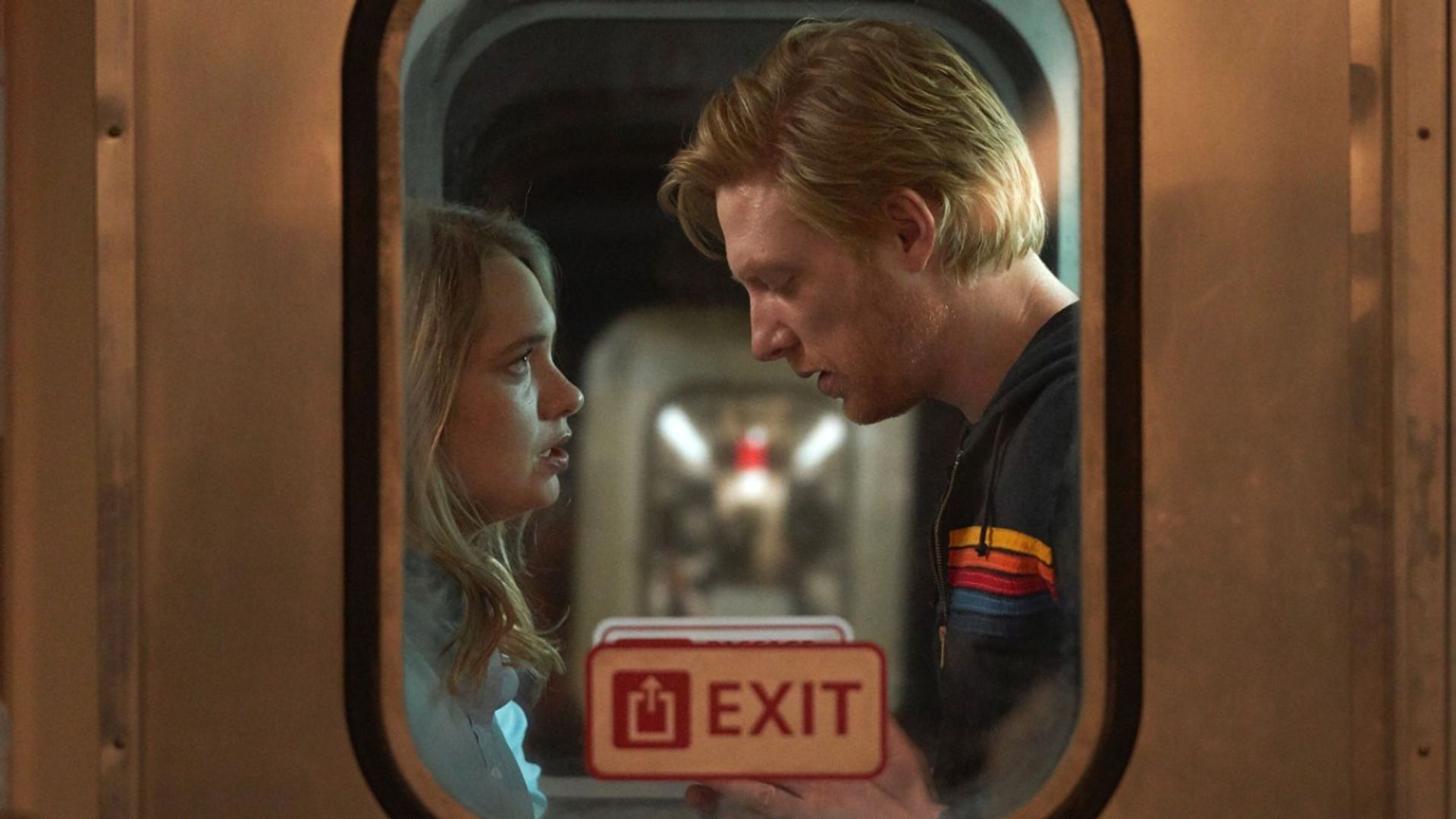 Domhnall Gleeson și Merritt Wever stau față în față în vestibulul trenului, filmați din afara ferestrei