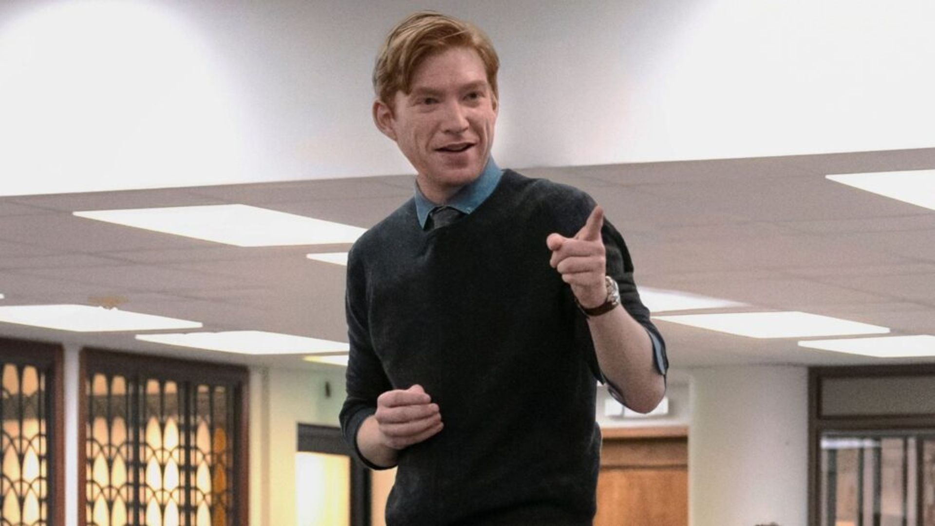 Domhnall Gleeson într-un pulover albastru închis stând pe o masă și arătând cu degetul