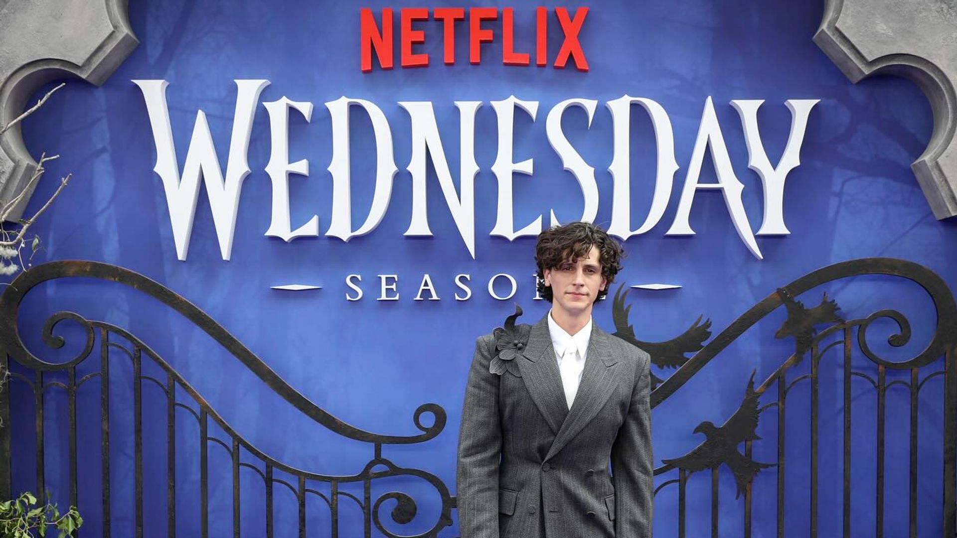 Owen Painter în costum stând în fața unui backdrop Netflix cu Wednesday
